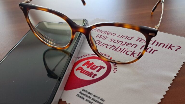 Brille auf einem Smartphone und einem Brillenputztuch mit MuT-Punkt-Logo